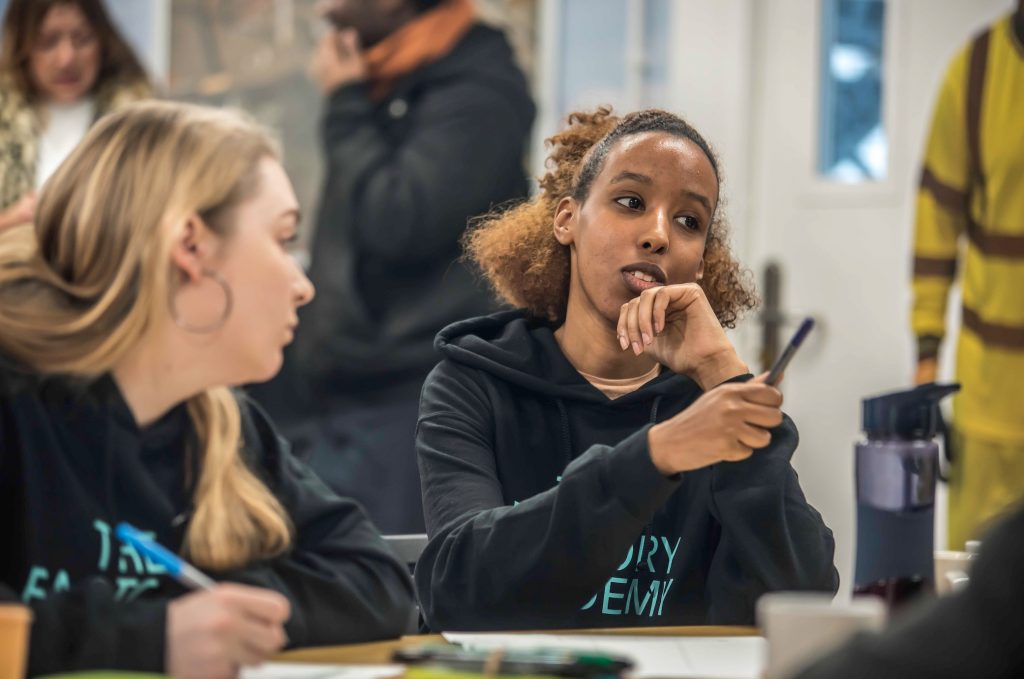 Two young people sat at a desk, with one actively taking part in the discussion signified by their hand gesture