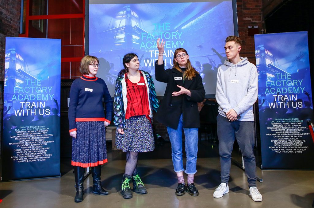 Four young stood people presenting in front of The Factory Academy banners
